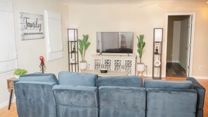 A living room with a blue sectional sofa, a flat-screen TV on a cabinet, and two potted plants.