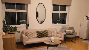 A living room with a beige sofa, a round coffee table, and a large mirror on the wall. There are two windows with curtains, a wooden cabinet, and a small plant on the left. - Southampton City View Apartment (Southampton)