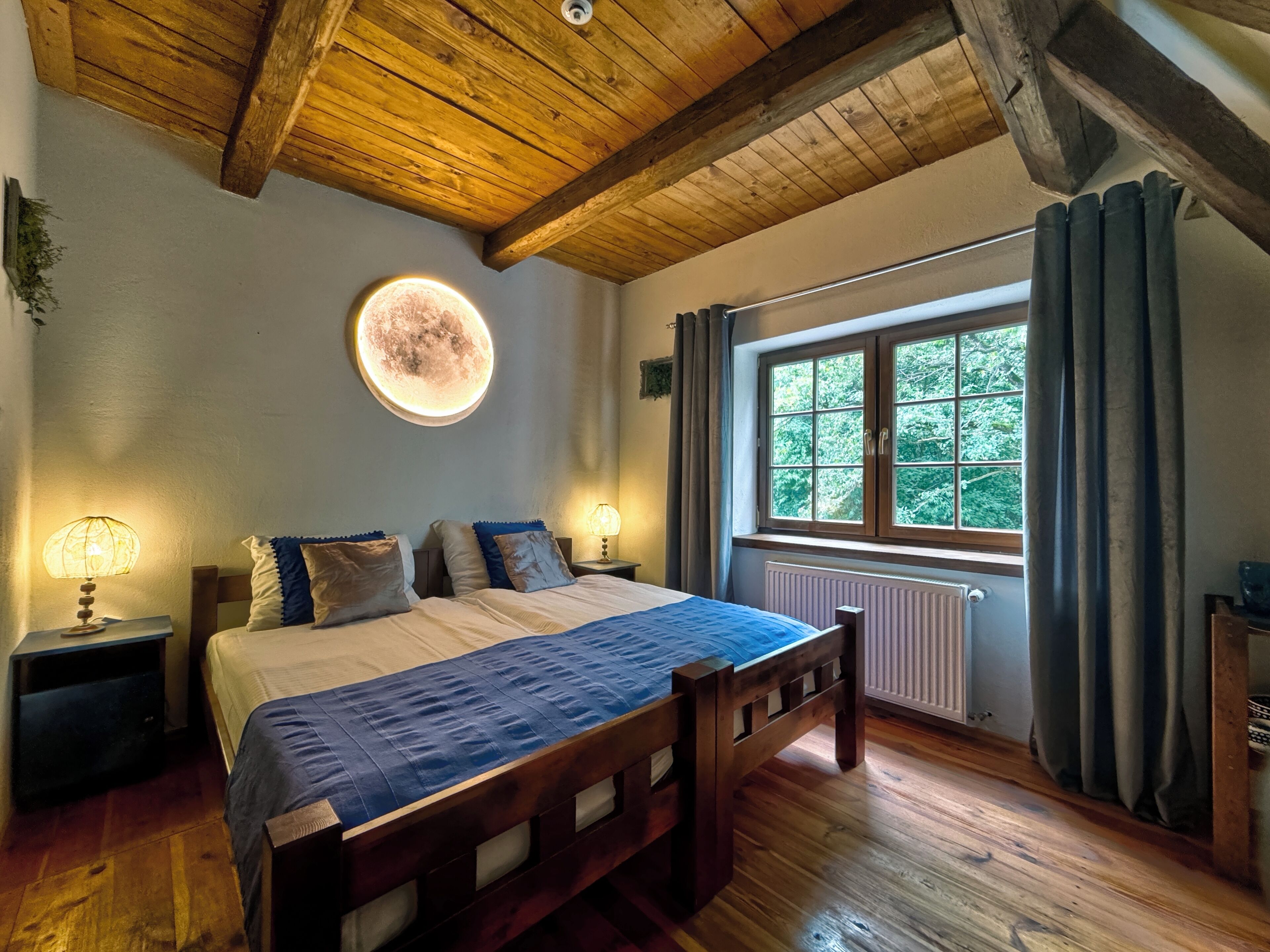 A bedroom with a wooden ceiling, a round wall light, a bed with a blue bedspread, two bedside tables with lamps, and a window with curtains.