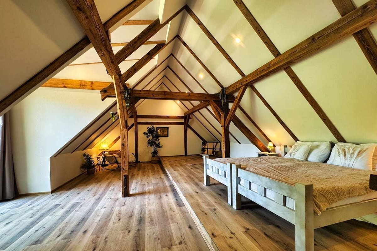 A room with a sloped ceiling, wooden beams, a bed, a desk, and a potted plant.