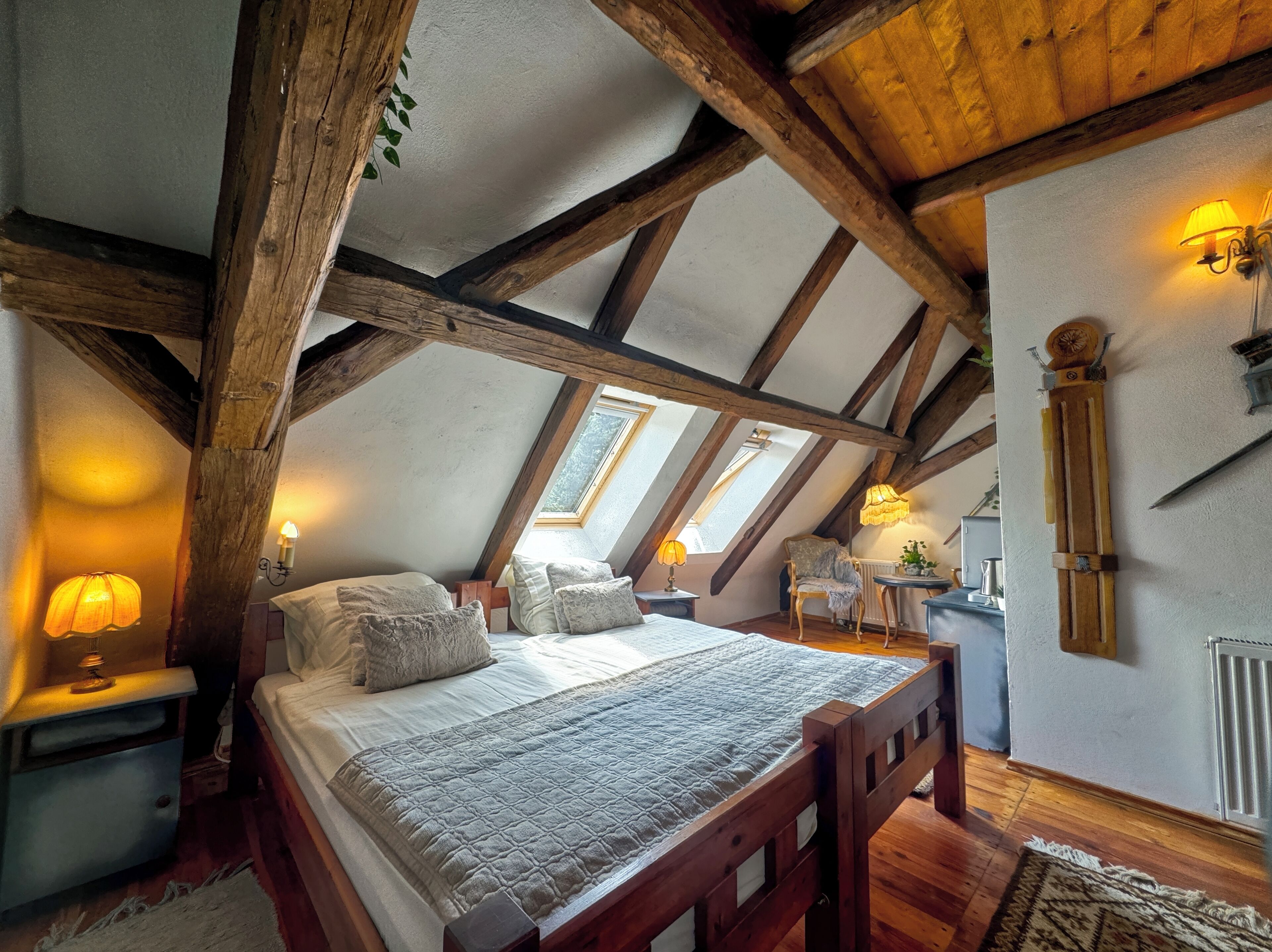 A bedroom with a sloped ceiling, wooden beams, a bed with pillows, bedside lamps, and a small table with a chair.