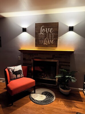 A cozy living room with a fireplace, a red chair, a decorative pillow, and a wall-mounted sign.