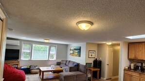 Ein großzügiger Wohnraum mit Ecksofa, Couchtisch und Fernseher. Es gibt eine Küchenzeile mit Holzschränken und Arbeitsplatte.