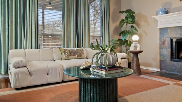Ein Wohnzimmer mit einer Couch, einem Glastisch, einem Kamin und einem großen Fenster mit Vorhängen.