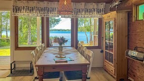A dining area with a wooden table and chairs, a vase with flowers, and a view of a lake through large windows. - Awesome home in Hultsfred with sauna (Hultsfred)