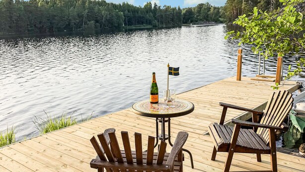 A wooden deck with a table and chairs overlooking a lake, a bottle of champagne, and a small Swedish flag. - Awesome home in Hultsfred with sauna (Hultsfred)