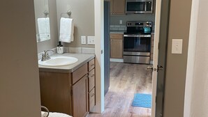 A bathroom with a toilet, sink, and mirror. The room has wooden flooring and a door leading to a kitchen with a microwave and oven. - Convenient and bright Cross Trail Vacation Getaway! (Sitka)