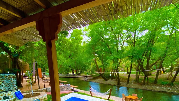 Un área de piscina con muelle de madera, una mesa y sillas, rodeada de exuberante vegetación y un río.