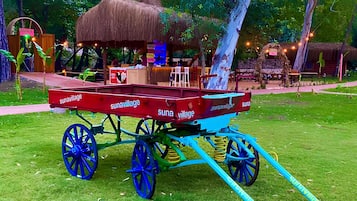 A traditional thatched-roof carriage with blue wheels and a canopy, parked on a grassy area with trees in the background.