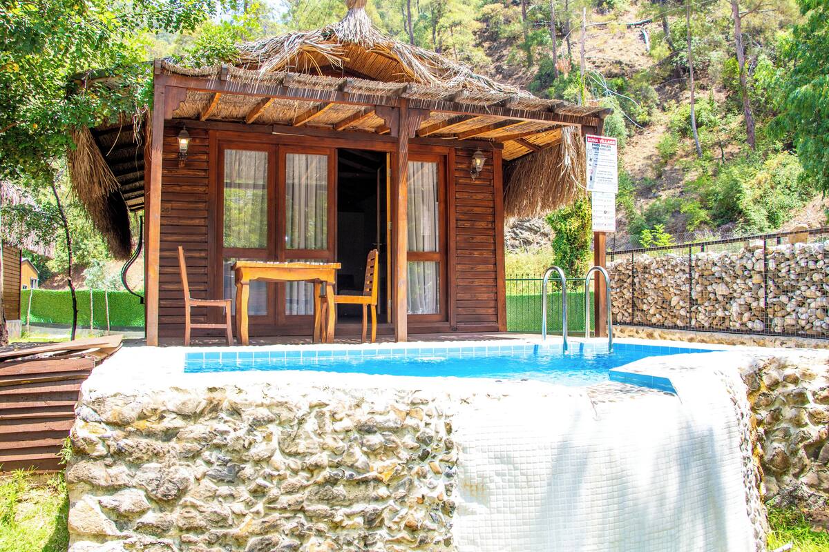 Una cabaña de madera con techo de paja, piscina y un muro de piedra.