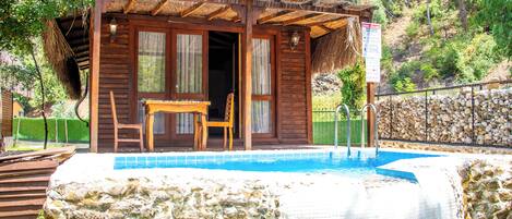A wooden cabin with a thatched roof, a pool, and a stone wall.