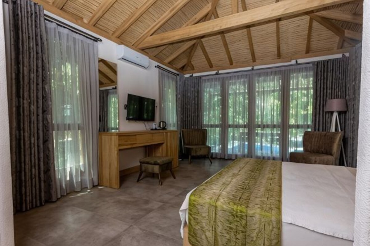 Una habitación con cama, escritorio, silla, televisión y ventanas amplias.