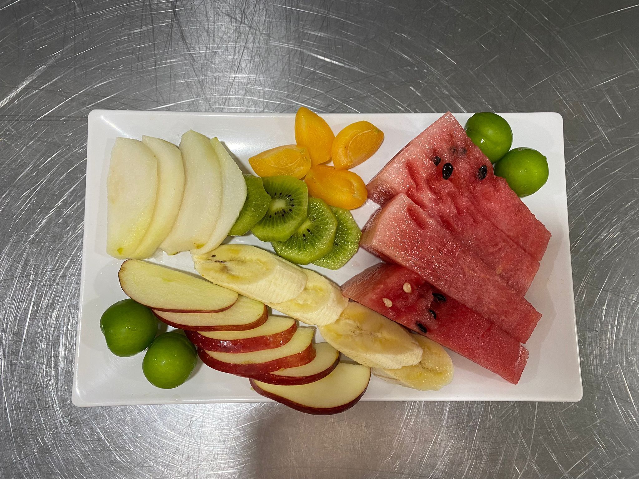 Un plato blanco rectangular con varias frutas, incluyendo rodajas de manzana, plátano, kiwi, sandía y limones.