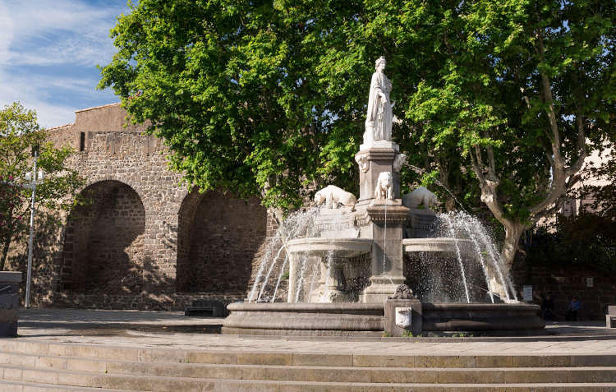 Une fontaine en pierre avec des jets d’eau, entourée d’une zone pavée et d’un mur en pierre orné d’arcades.