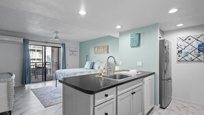 A modern kitchen with a black countertop, white cabinets, and a stainless steel refrigerator. There is a dining area with a table and chairs, and a balcony with blue curtains.