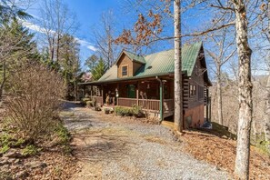 Une cabane en rondins avec un toit végétalisé, une véranda et une allée de gravier, le tout entouré d’arbres.