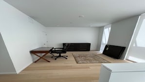 A modern, sparsely furnished room with a wooden floor, a small desk, a black office chair, a black entertainment unit, and a large window.