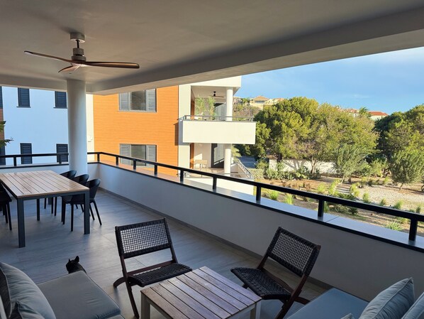 Un balcon avec une table et des chaises en bois, un ventilateur de plafond et une vue sur les bâtiments et les arbres.