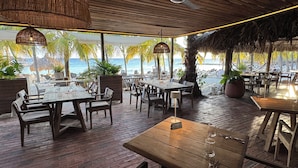 An outdoor dining area with wooden tables and chairs, a thatched roof, and a view of the beach.