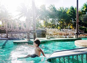 Zona de piscina con un niño jugando, rodeada de palmeras y un muro de piedra.