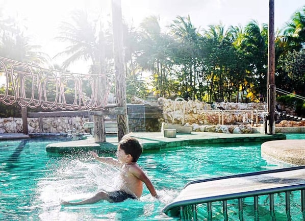 A pool area with a child playing, surrounded by palm trees and a stone wall.