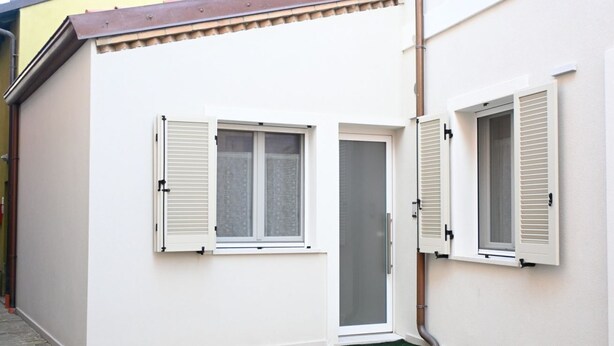Ein kleines, weißes Gebäude mit zwei Fenstern, eines davon mit Fensterläden, und einer Tür mit einer Glasscheibe.