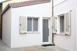 Un petit bâtiment blanc, doté de deux fenêtres, dont une avec des volets, et d’une porte avec un panneau de verre.