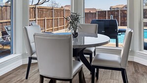 Un coin repas avec une table ronde, six chaises et un lustre. De grandes fenêtres en arc offrent une vue sur un jardin avec une clôture en bois et un barbecue.