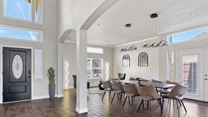 Un vaste espace dédié à la salle à manger, avec une grande table et des chaises, une porte noire agrémentée d’un panneau de verre décoratif et une plante en pot.