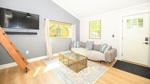 Una sala de estar con un sofá, una mesa de centro, un televisor montado en la pared y una ventana con vista a una casa.