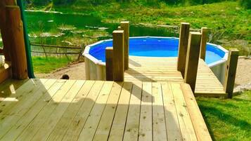 Una terraza de madera que lleva a un jacuzzi circular rodeado por una valla de madera, con un césped y árboles al fondo.