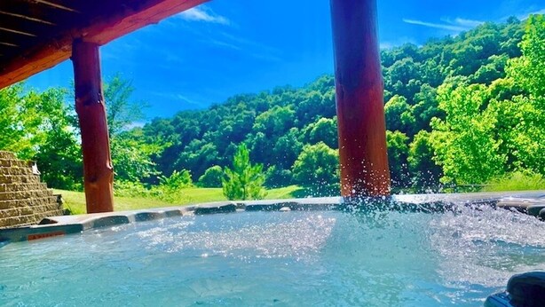 Un jacuzzi con chorros de agua, rodeado por una terraza de madera y una estructura de piedra, con un bosque verde y frondoso al fondo.