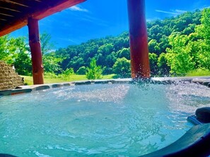 Un jacuzzi con chorros de agua, rodeado por una terraza de madera y una estructura de piedra, con un bosque verde y frondoso al fondo.