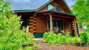 Una cabaña de troncos con techo verde, un porche con mobiliario exterior y un pequeño jardín.