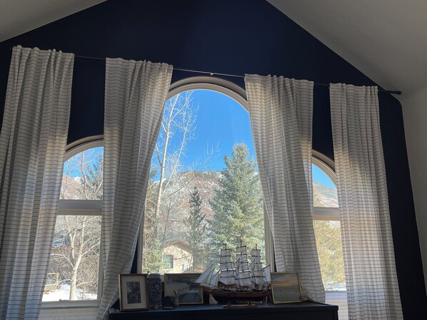 A room with a large arched window, white curtains, and a small table with a ship model and framed pictures.