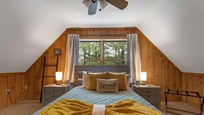 Une chambre mansardée chaleureuse avec un plafond en pente, des murs en bois, une grande fenêtre avec des rideaux, un lit avec une courtepointe et des oreillers, des lampes de chevet et une petite horloge au mur.