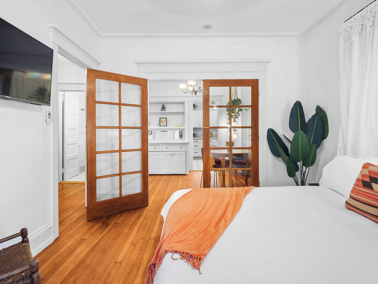 A bedroom with a large bed, a wooden floor, a white wall, and a view into a kitchen area.