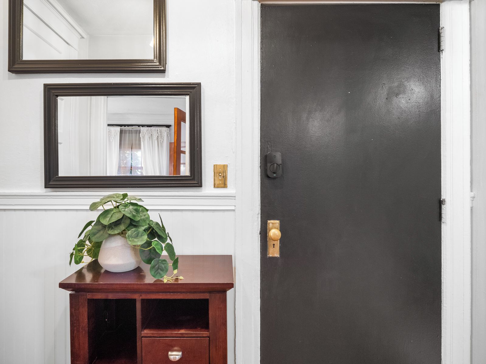 A room with a dark door, a mirror above a wooden console table with a potted plant, and a light switch on the wall.