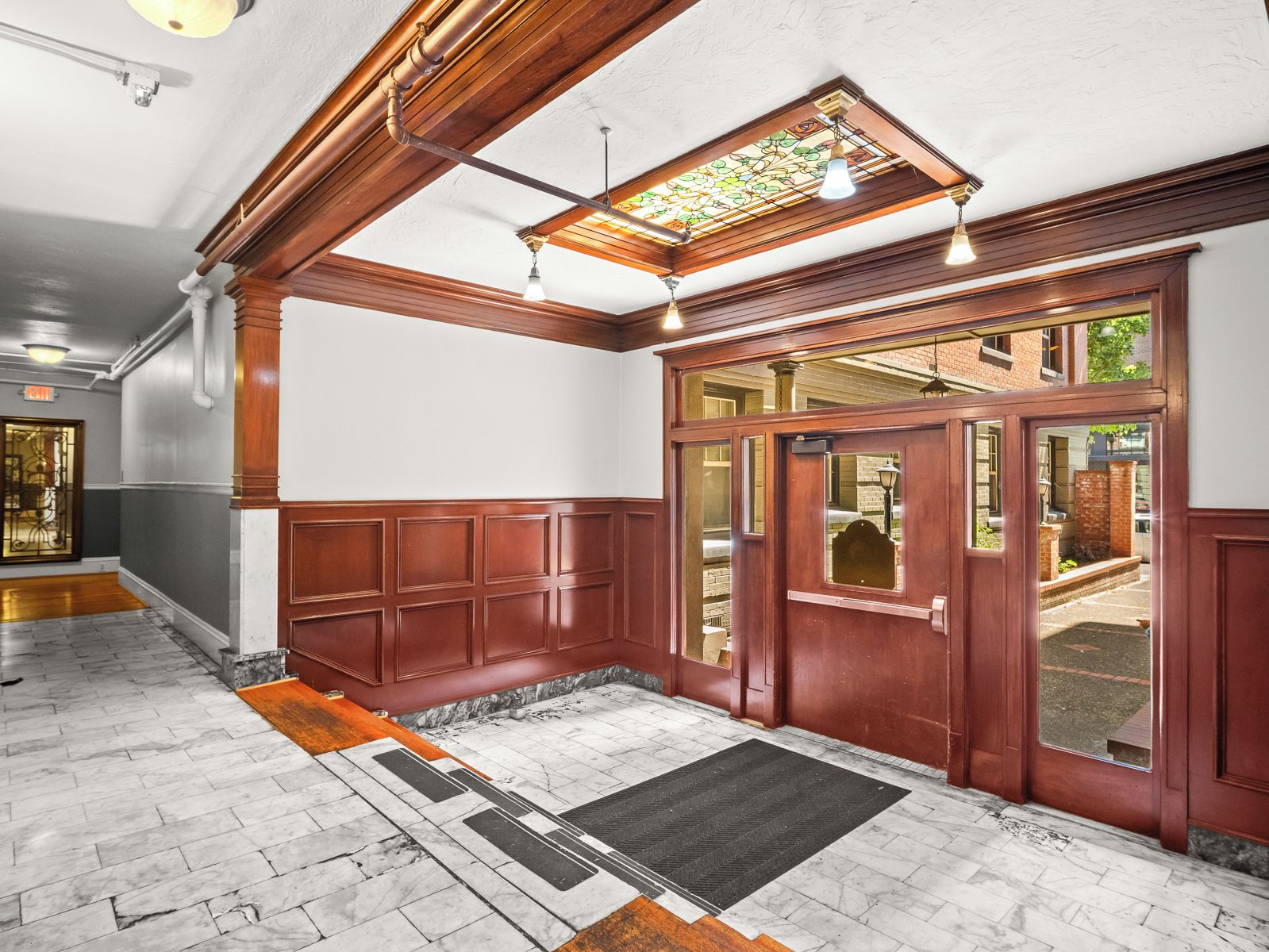 A hallway with a wooden door, a black mat at the entrance, and a stained glass window above.