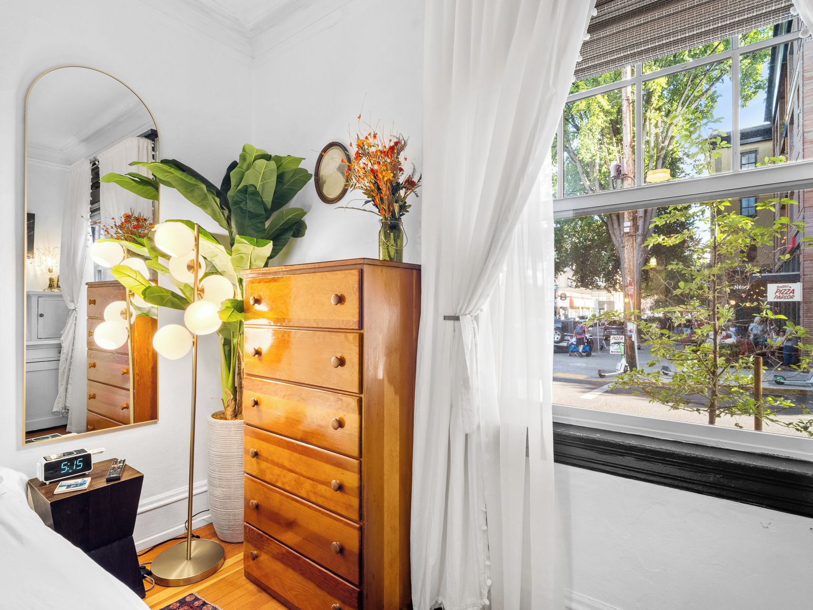 A room with a wooden dresser, a mirror, a clock, and a window with a view of the street.