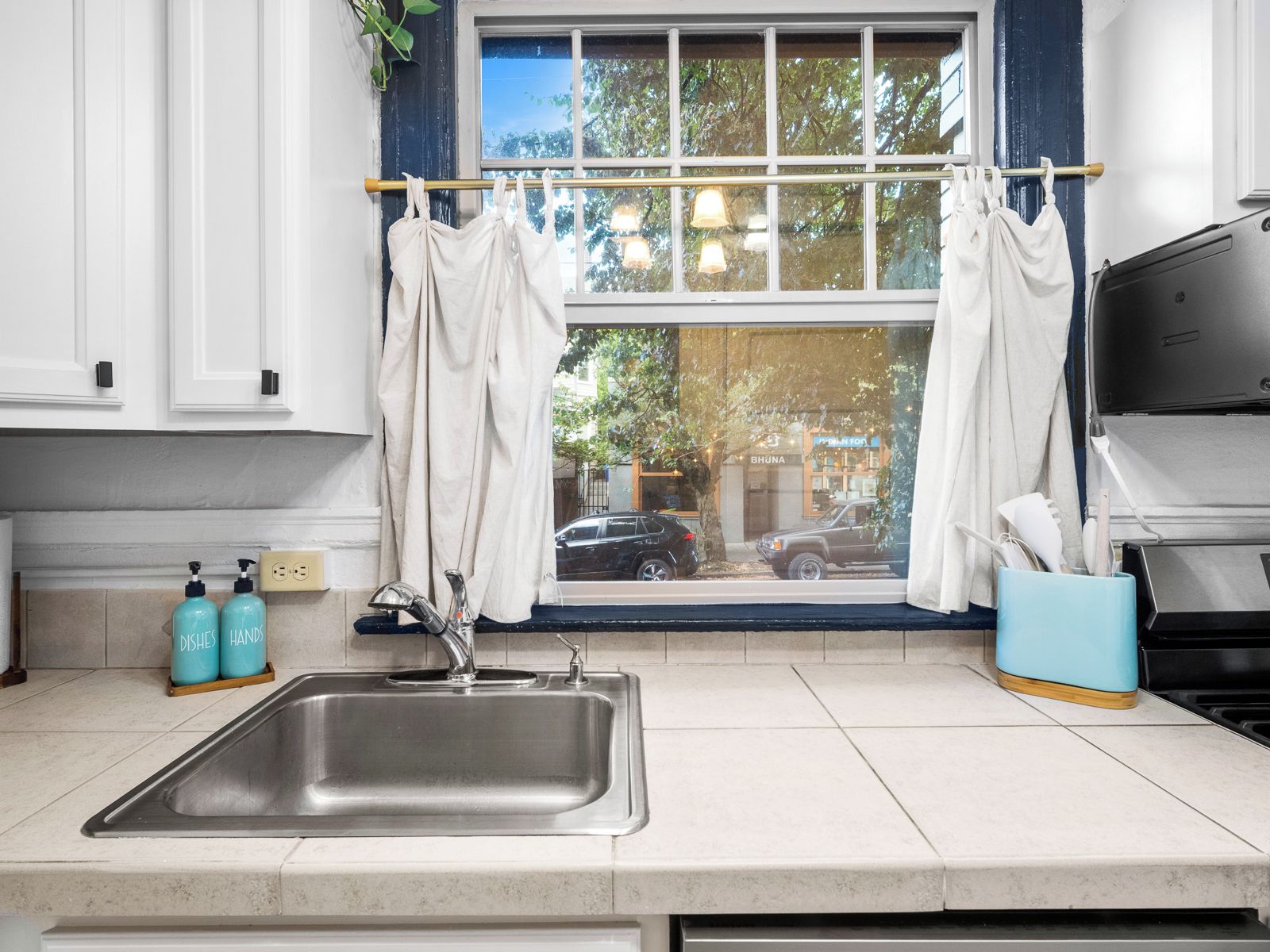 A kitchen with a stainless steel sink, a window with curtains, and a view of the street outside.
