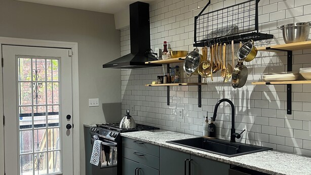 A modern kitchen with grey cabinets, a black stove, and a black sink. There is a black pot rack above the sink and a wooden door with a window. - Stylish Townhouse by Carrol Park, M&T Bank Stadium (Baltimore)