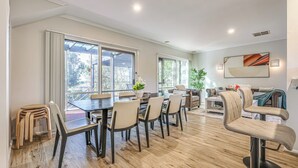 Un comedor amplio con una mesa y sillas de madera, ventanas grandes con vistas al exterior, y una sala de estar moderna con un sofá y plantas decorativas.