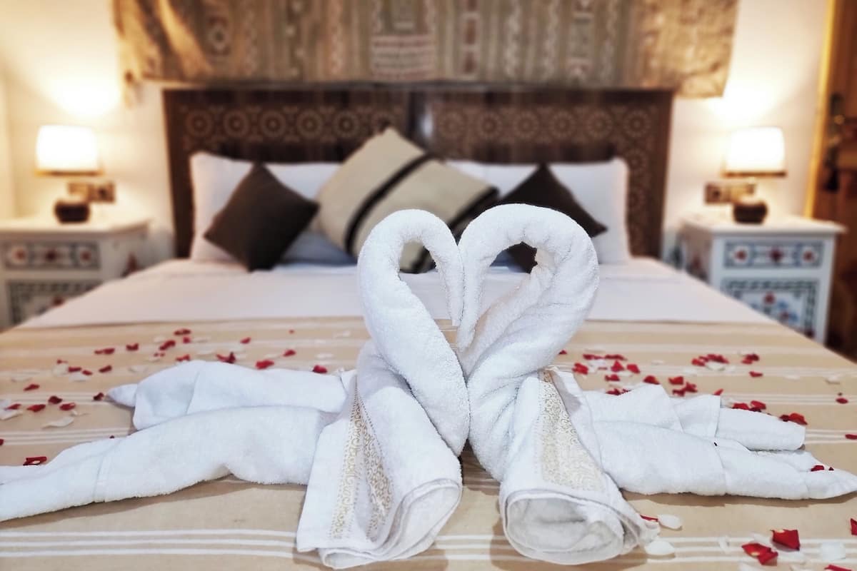 Una habitación de hotel con una cama, dos mesitas de noche, un cabecero con un diseño estampado y una decoración con toallas en forma de corazón sobre la cama.