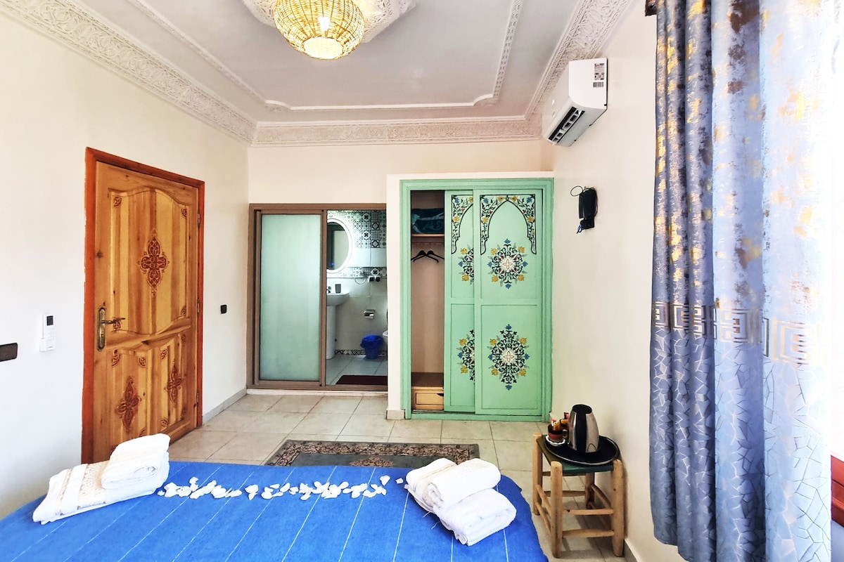 A hotel room with a bed, a wooden door, a green wardrobe, a bathroom with a mirror, and a ceiling light.