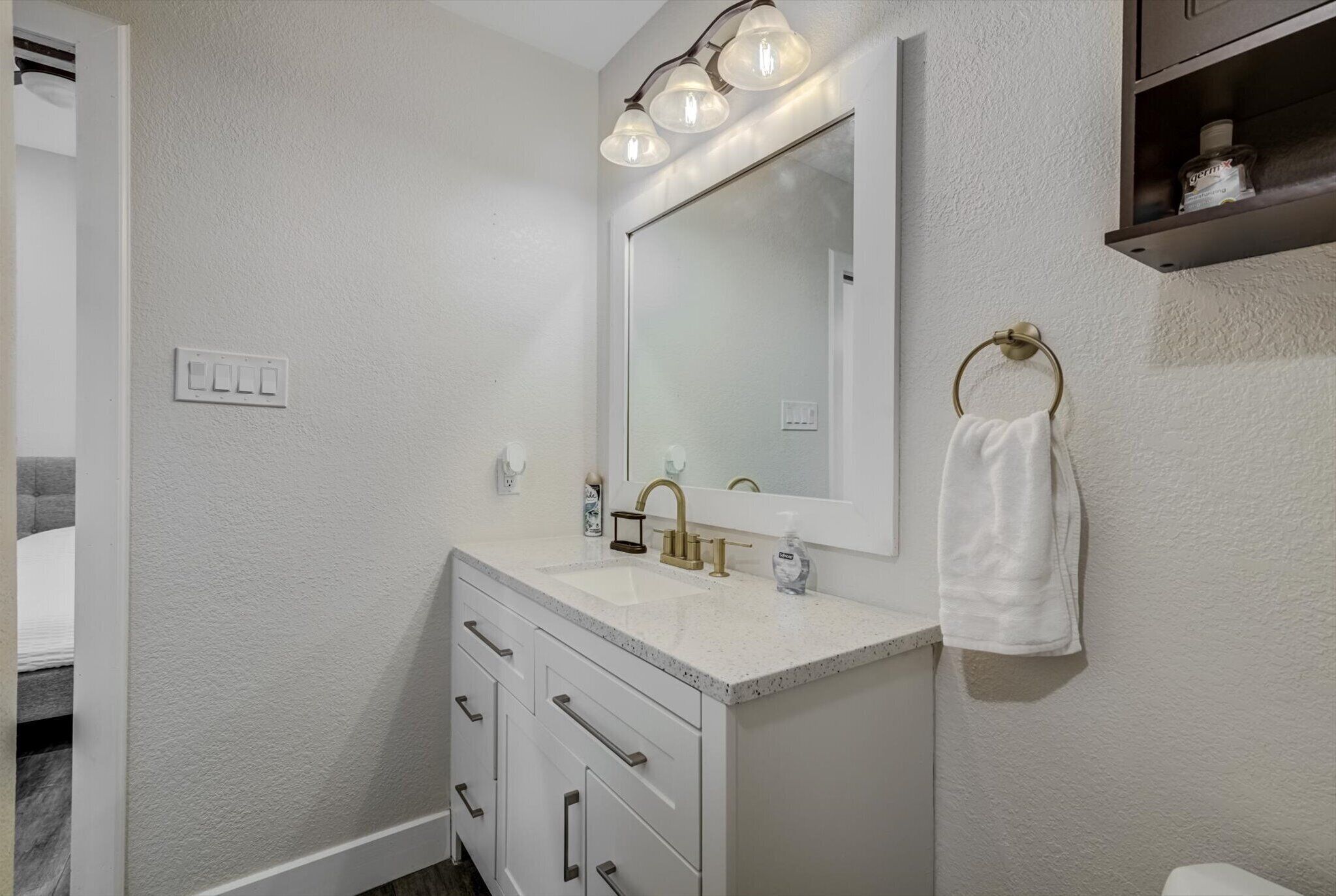 A bathroom with a white countertop, a gold faucet, and a mirror with three lights. There is a towel ring on the wall and a small shelf above the mirror.