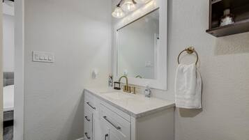 A bathroom with a white countertop, a gold faucet, and a mirror with three lights. There is a towel ring on the wall and a small shelf above the mirror.