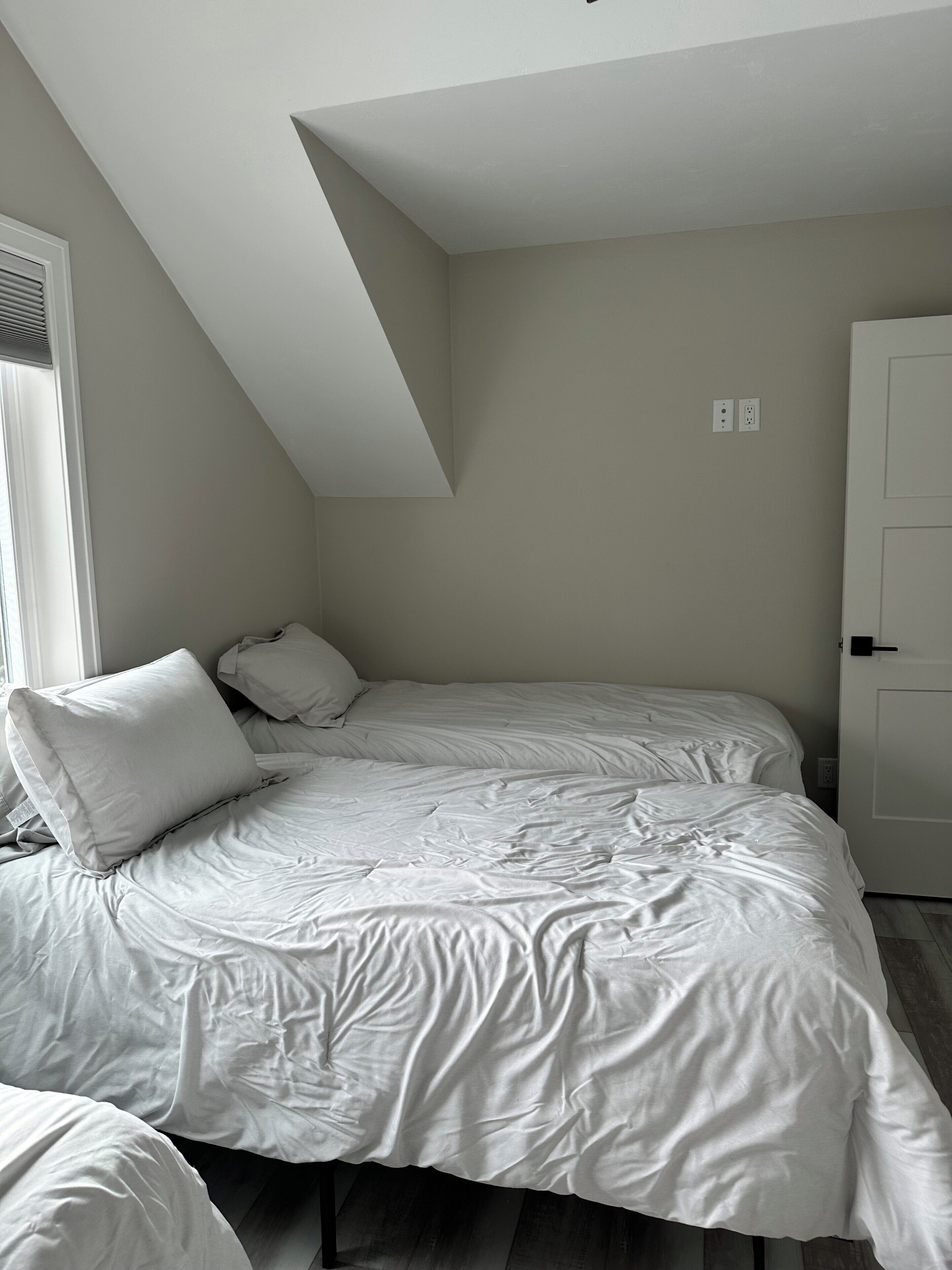 Ein Schlafzimmer mit schrägen Wänden, einem Bett mit weißer Bettwäsche, einem Fenster mit Jalousien und einer Tür.
