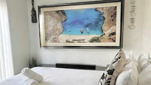 Ein Schlafzimmer mit einem großen, gerahmten Küstenbild, einem Bett mit weißer Bettwäsche und einer kleinen Pflanze.