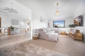 A modern living room with a white sofa, a dining area with a white table, and a large window with a view of the sea.